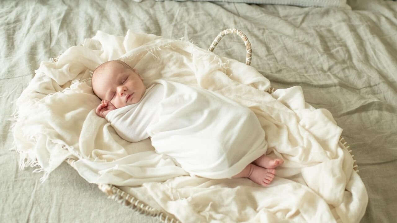 Newborn baby sleeps soundly in a woven basket, wrapped in a soft white blanket. A peaceful newborn session by Moments in Light Photography.