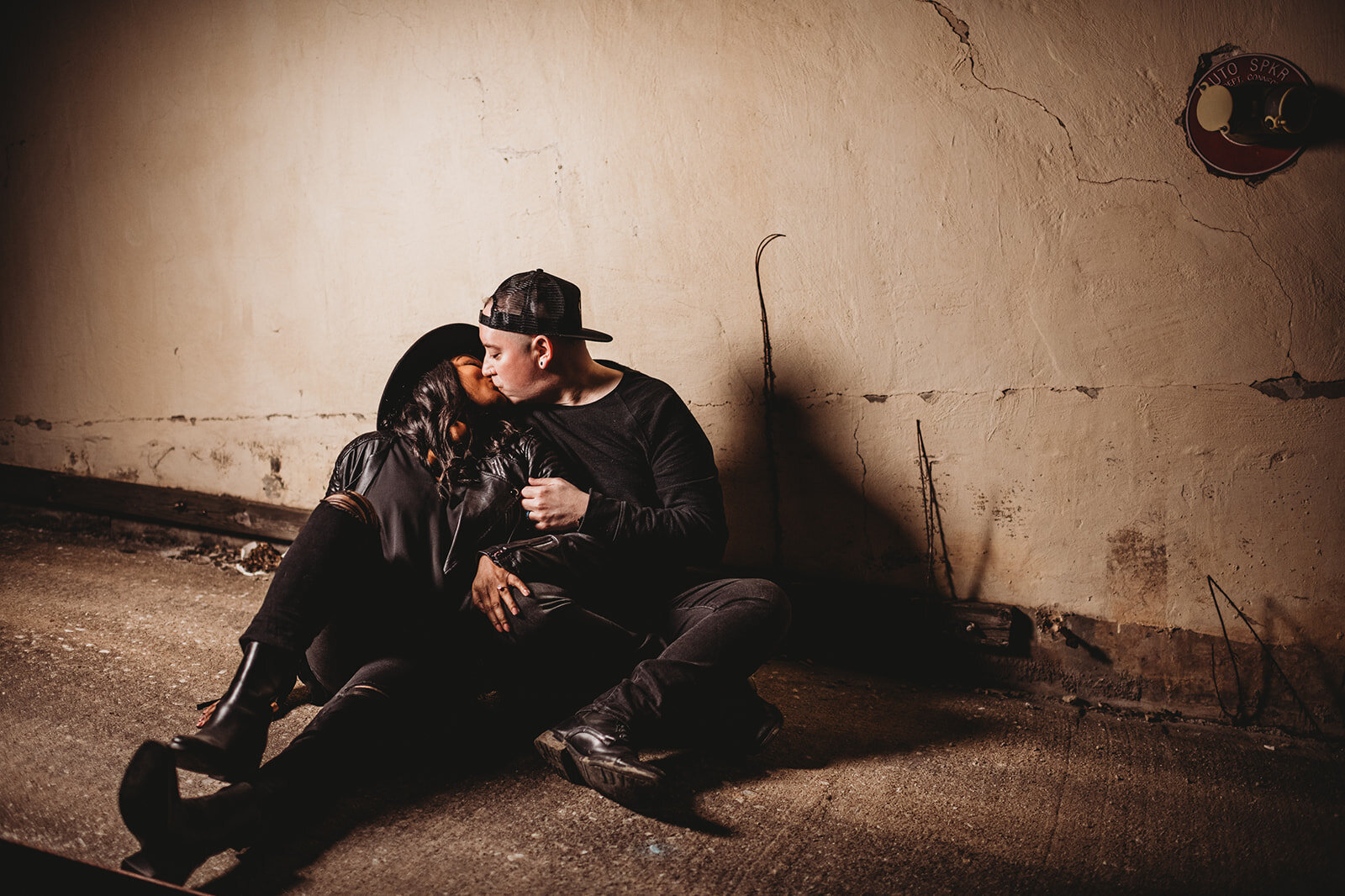 Outdoor engagement photos with man and woman sitting on a sidewalk leaning against a concrete wall as they kiss one another captured by Baltimore photographers