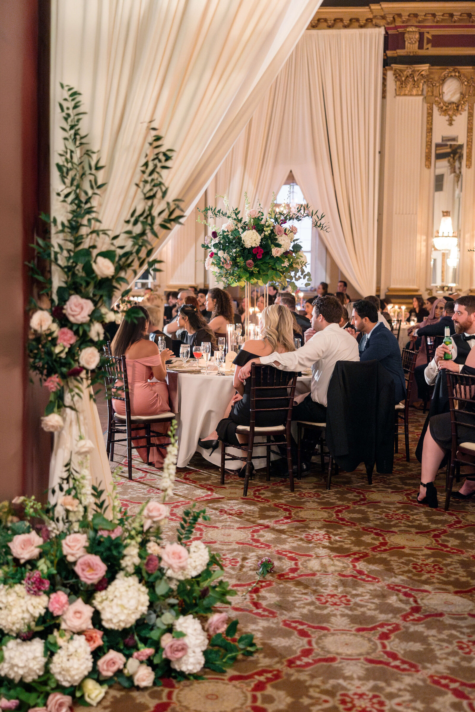 A formal event with elegantly dressed people seated around tables decorated with large floral centerpieces. The room features ornate decor with white drapes and a patterned carpet. Ambient lighting adds a warm atmosphere.