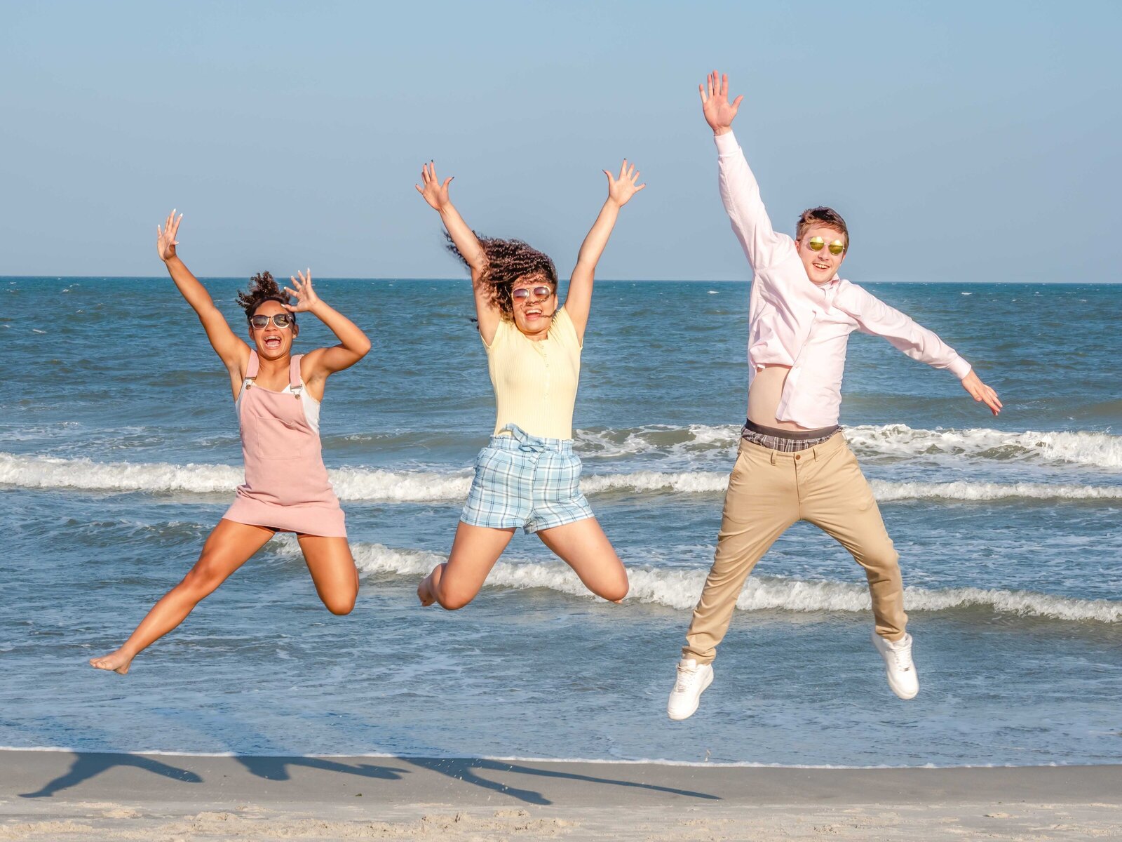 Three friends joyfully jump in the air at Carolina Beach, wearing casual summer outfits and sunglasses. The ocean waves and clear blue sky provide a lively backdrop. This image showcases affordable family photography in Wilmington, NC, perfect for capturing fun, energetic moments.