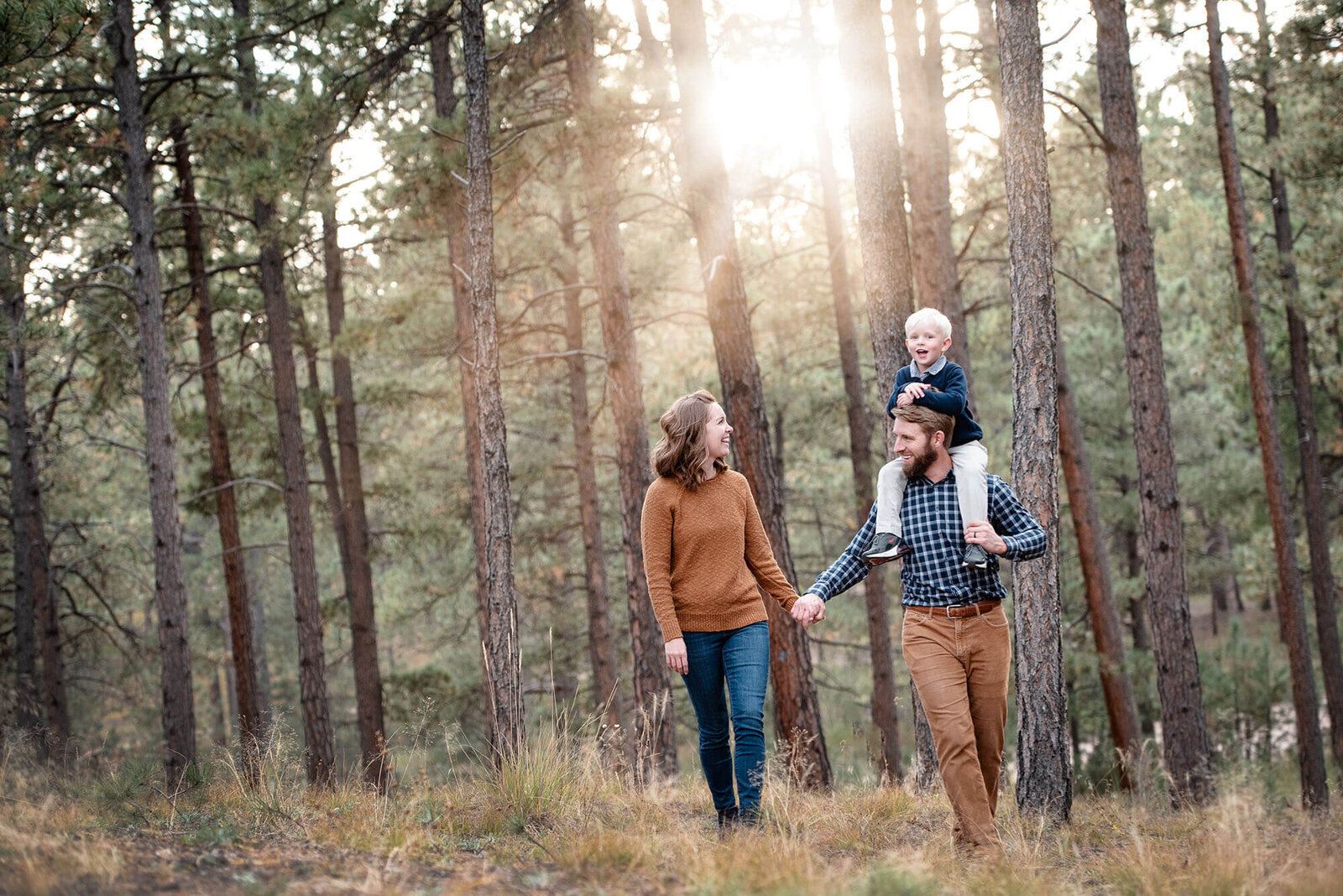 A mom and dad hike through a forest at sunset with a toddler on dad's shoulders