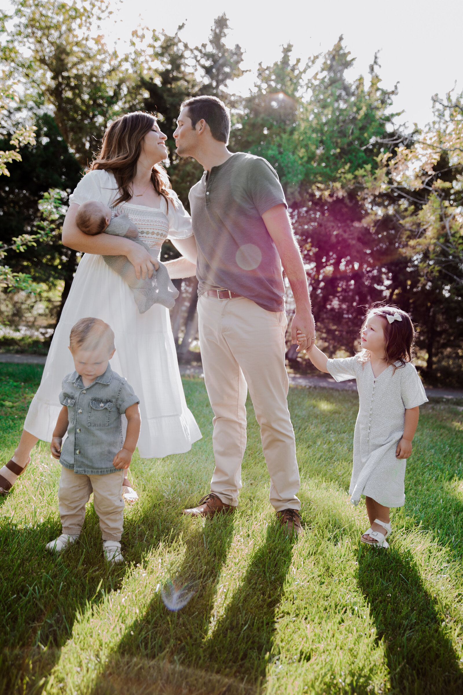 outdoor summer family photos with Idaho Falls Family photographer as the mother and father kiss while the mother holds their newborn baby and their small children run around their feet