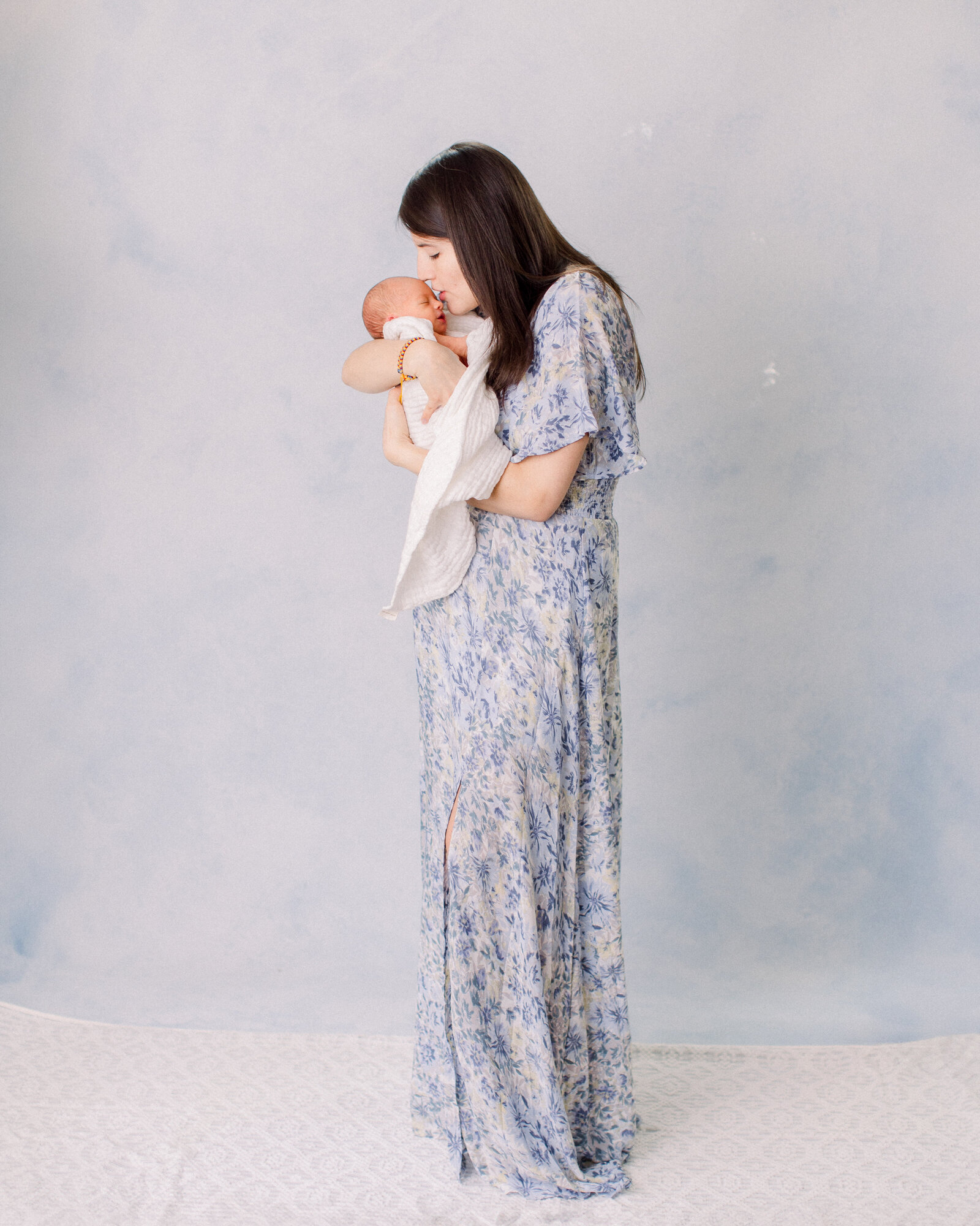 New mom in blue and white floral gown cradling her sleeping her baby in a studio with a blue hand painted background by NH newborn photographer Fieldstone Studio.