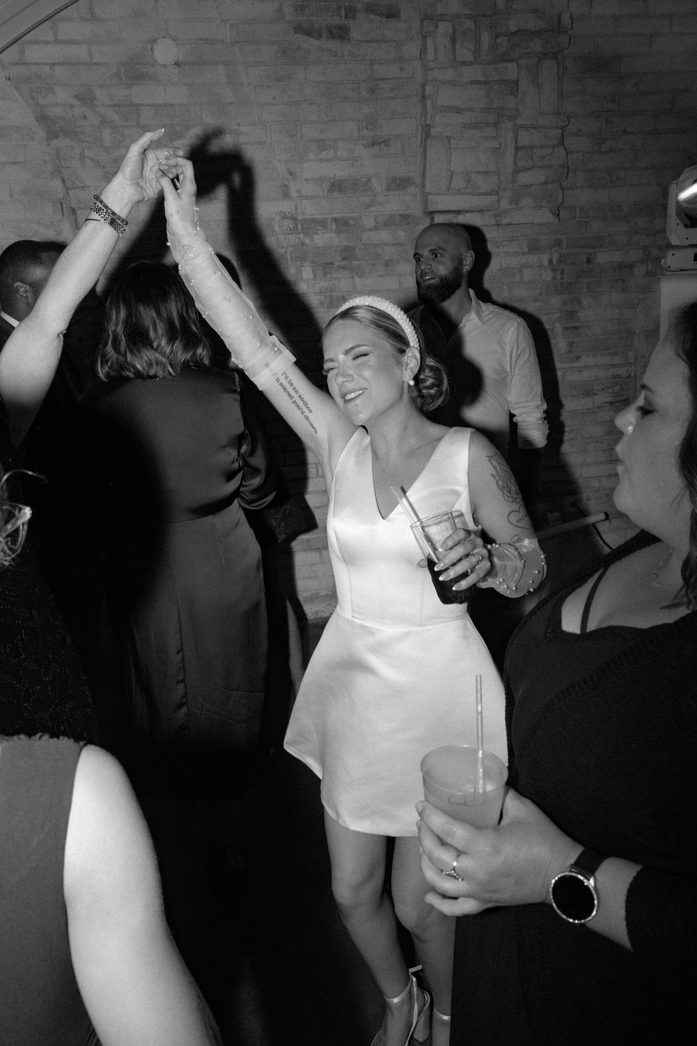 a bride dances joyfully with guests at a wedding party, smiling and holding a drink in one hand in a lively black and white scene.