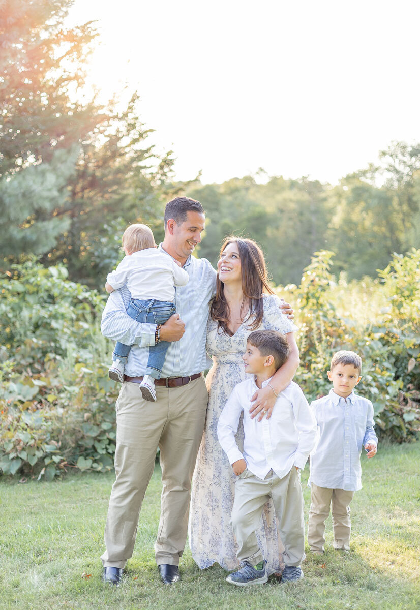 Smiling family of five during a golden hour photo session in Connecticut, with soft natural light, candid laughter, and lush greenery in the background—perfect for modern family lifestyle photography.