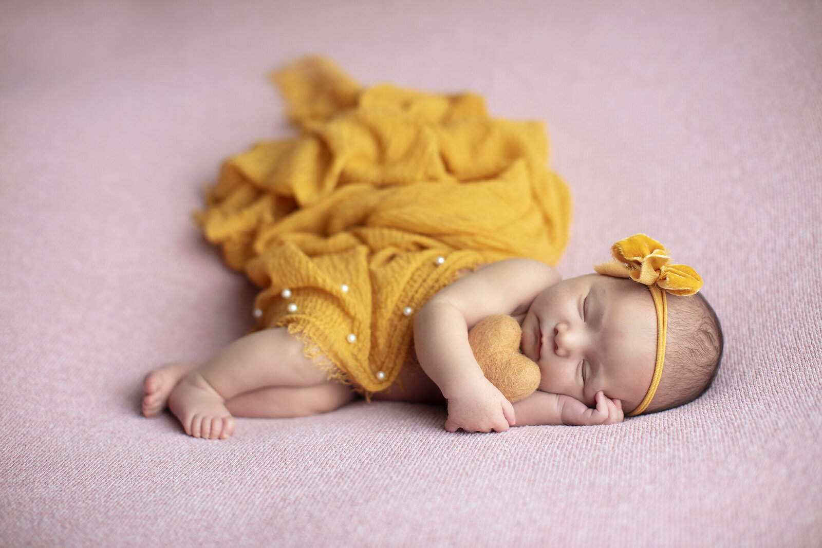 Newborn girl holds yellowhear on pink fabric at newborn photo shoot