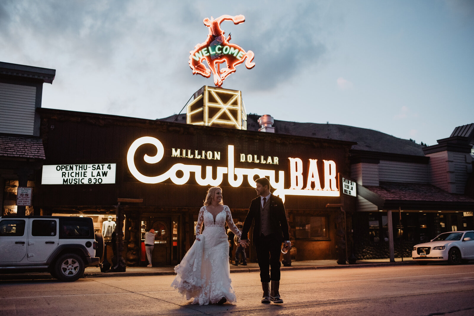 Cowboy Bar in Jackson with bride and groom holdin ghands and walking across the road as the sun sets behind the building and the lights shine from the bar onto the street
