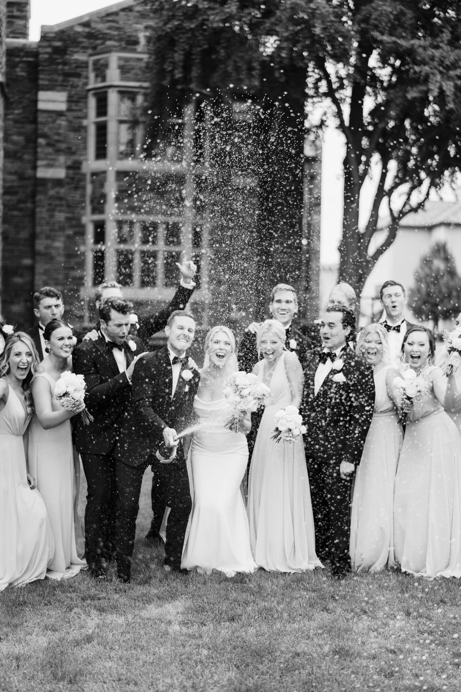 Groom pops open a bottle of champagne while the bride and the wedding party cheer