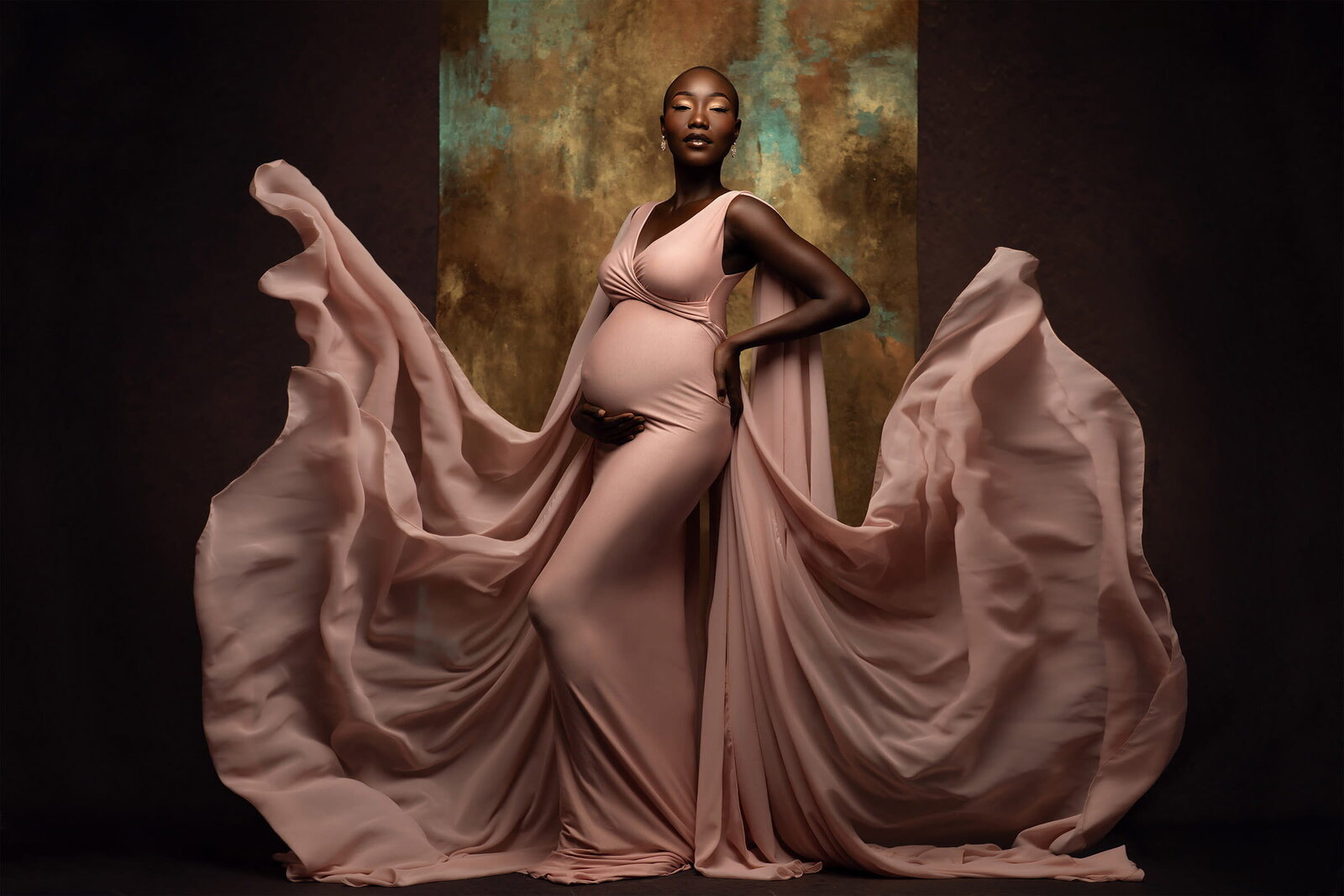A stunning pregnant woman poses gracefully in a blush pink gown with dramatic flowing fabric surrounding her. Her hand rests on her belly as she stands against a gold and teal textured backdrop. Photo by DFW Maternity Photographer Lindsay Walden.