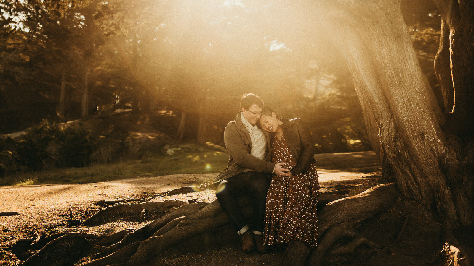 Maternity couple sits on tree stump and snuggles pregnancy belly with golden light streaming in