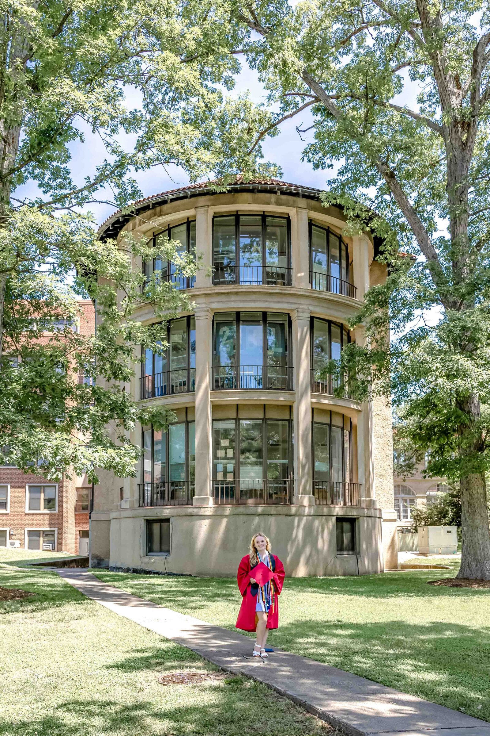 A NCSSM graduating senior in a red graduation gown holding a diploma, walking in front of a historic North Carolina School of Science and Math building surrounded by trees in Durham, North Carolina. This image captures academic achievement in a picturesque setting, perfect for memorable senior portraits.