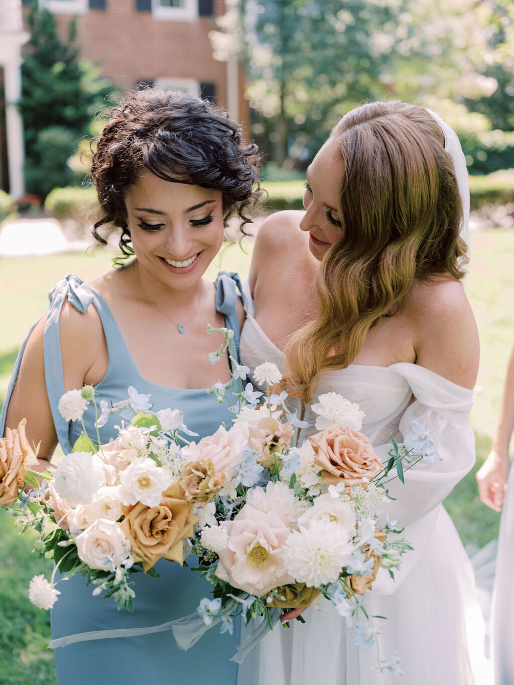 Bride and bridesmaid in blue dresses holding bouquets of peach garden rose, white dahlia, toffee roses, white hydrangea, light blue delphinium and brown lisianthus.
