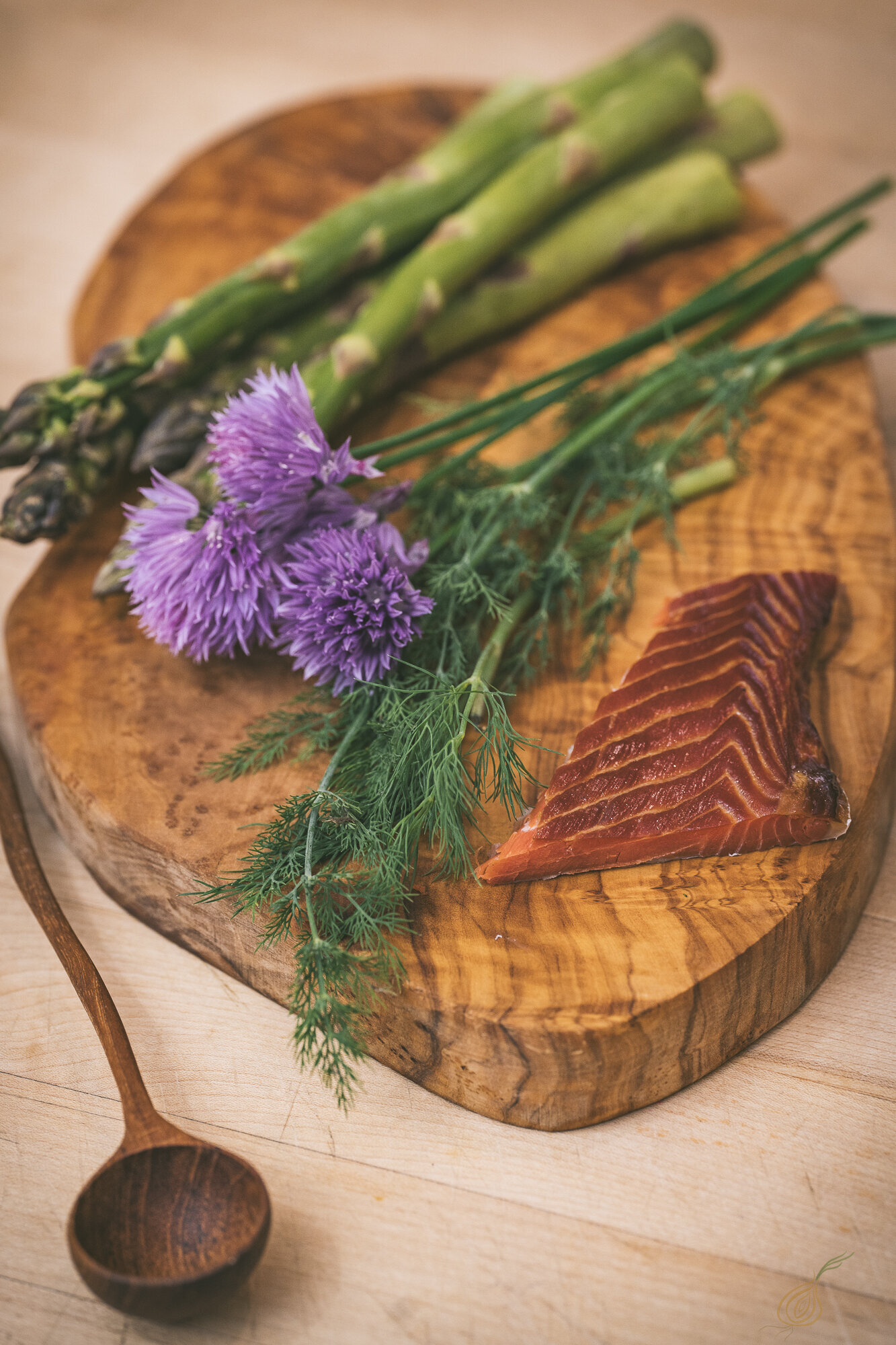 A wooden cutting board displays smoked salmon, fresh asparagus, and vibrant purple flowers, creating an elegant culinary scene.