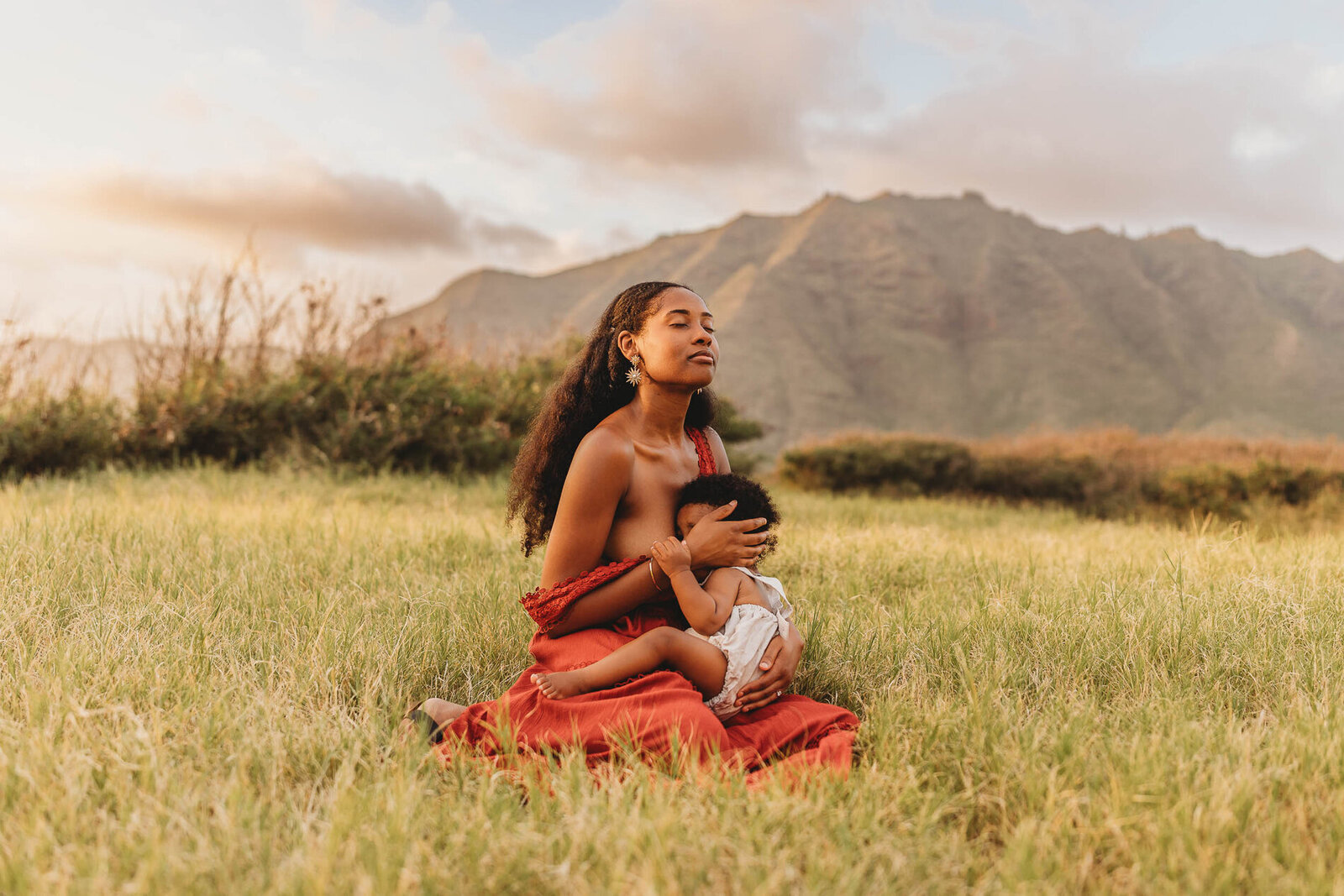 Mother breastfeeding her baby in a red dress against the mountains of Oahu — heartfelt motherhood and family photography by Leslie Carbajal