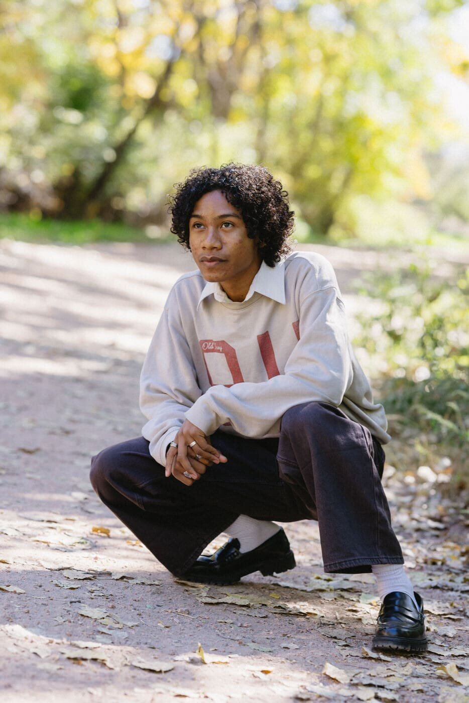 A high school senior boy crouches on a dirt path and looks off to the side.