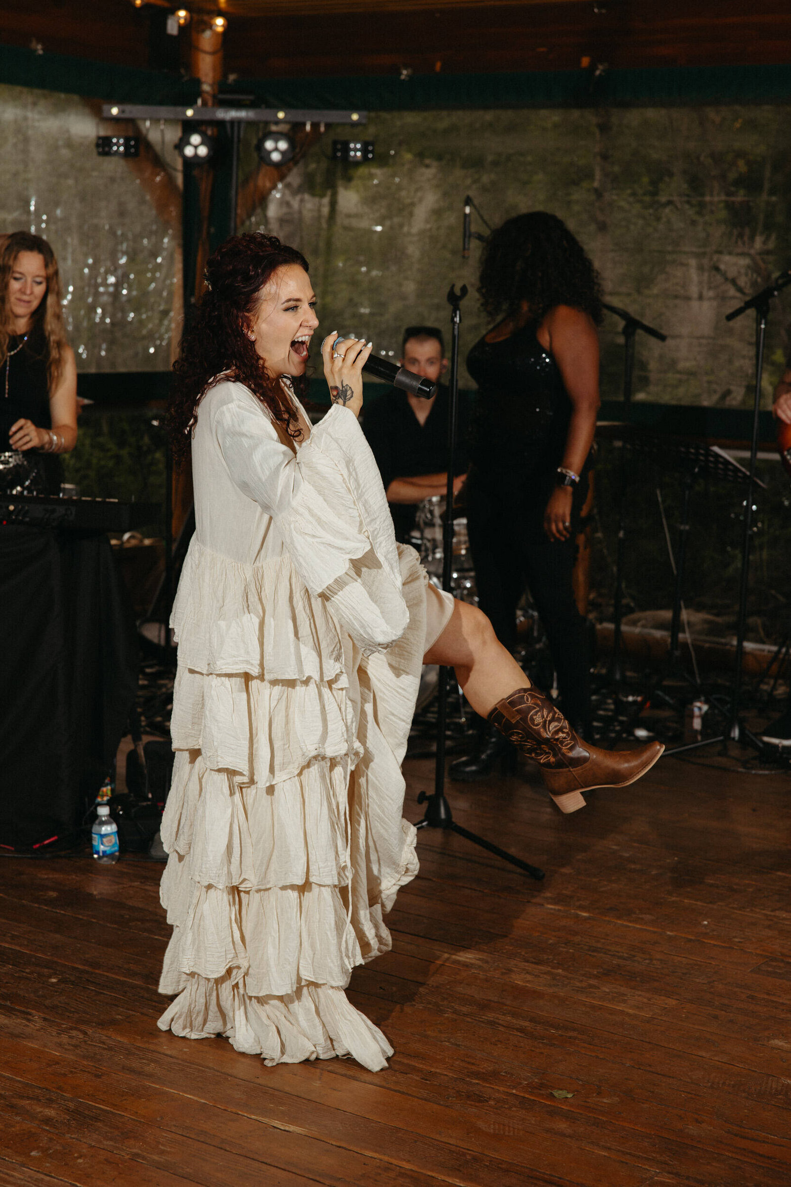 Bride wearing a festival style gown and cowboy boots cheers in the microphone while kicking up her foot on the dance floor