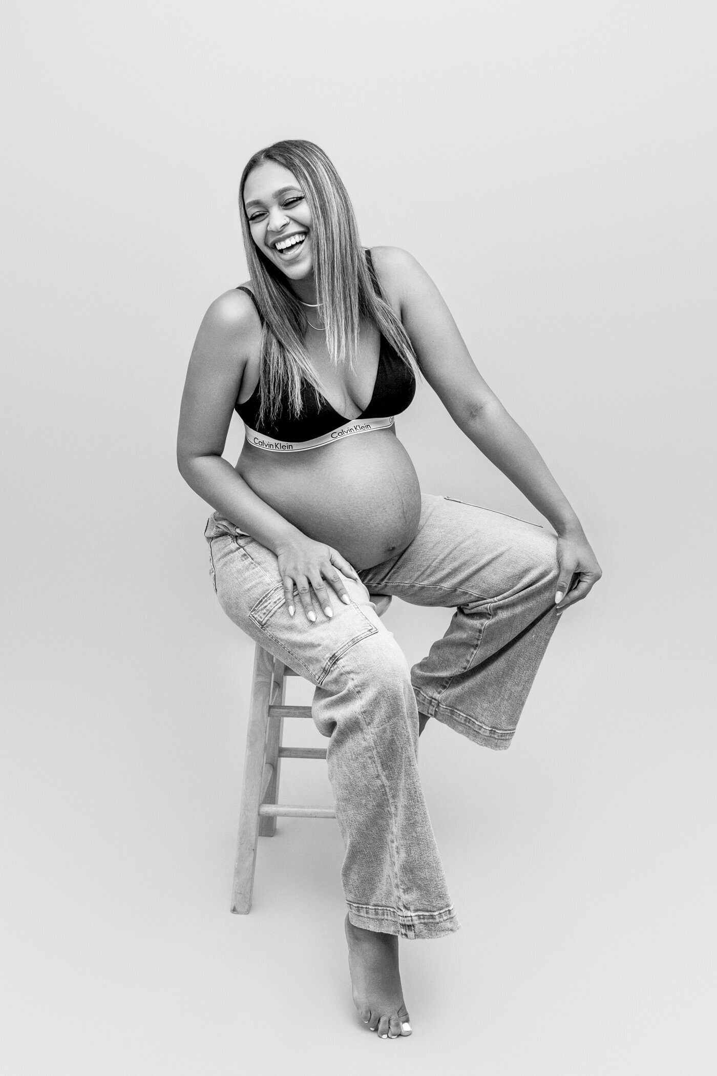 A joyful pregnant woman sits on a wooden stool, dressed casually in a black sports bra and loose-fitting jeans. She laughs heartily, tilting her head back with her hands resting on her knees, one of her legs crossed over the other. The black-and-white image captures her relaxed, carefree energy and the natural beauty of her pregnancy.