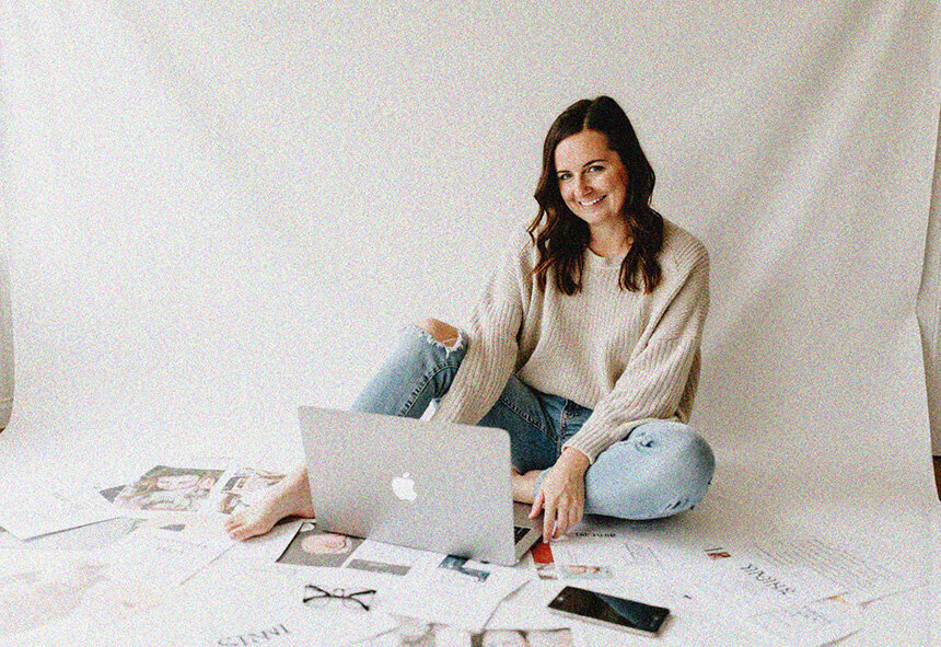 A woman named Jordin, a Showit web designer, sits on a white floor working on her laptop.