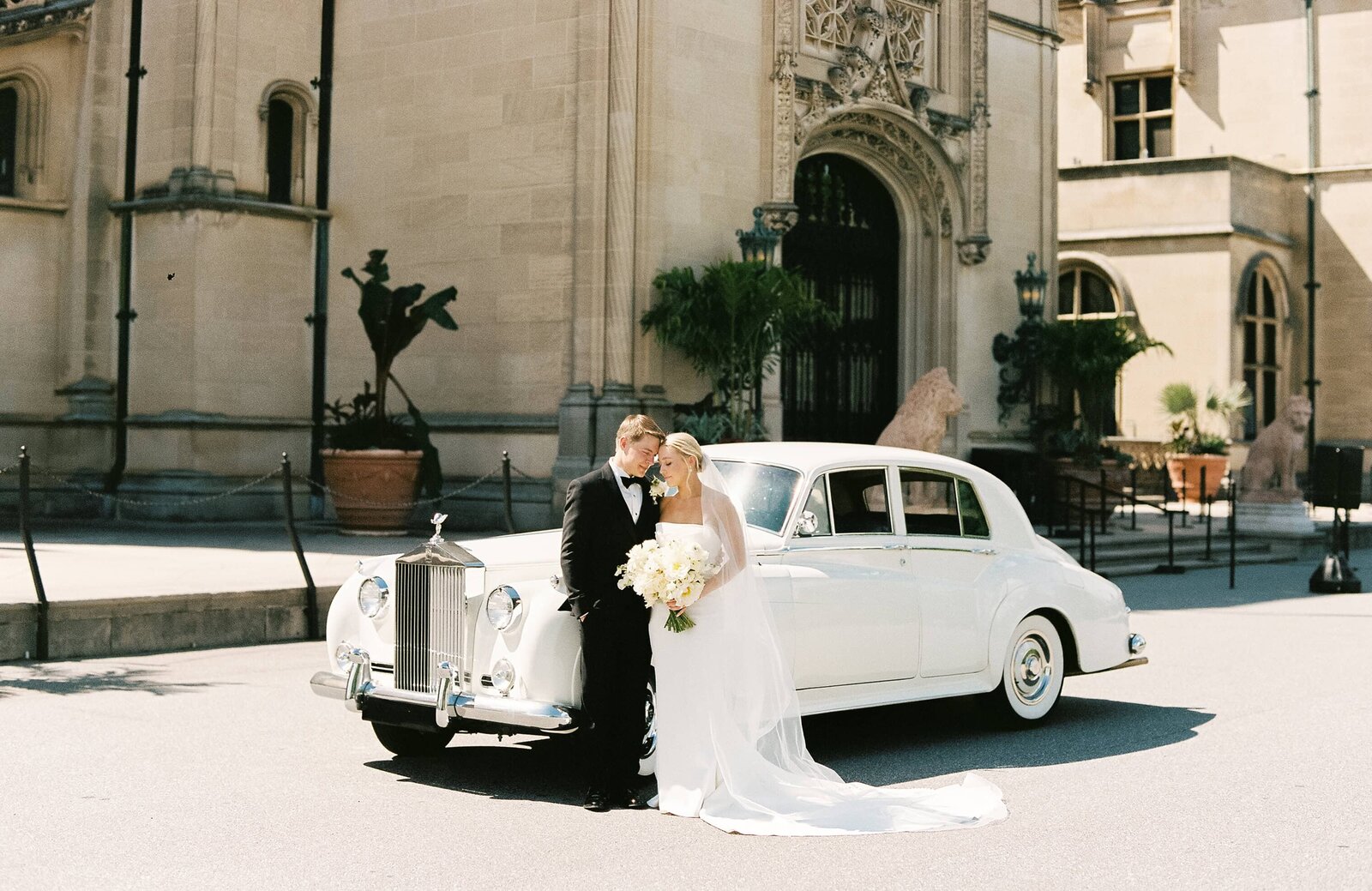 A bride and groom pose in front of an old car parked in front of the Biltmore Estate in Asheville North Carolina