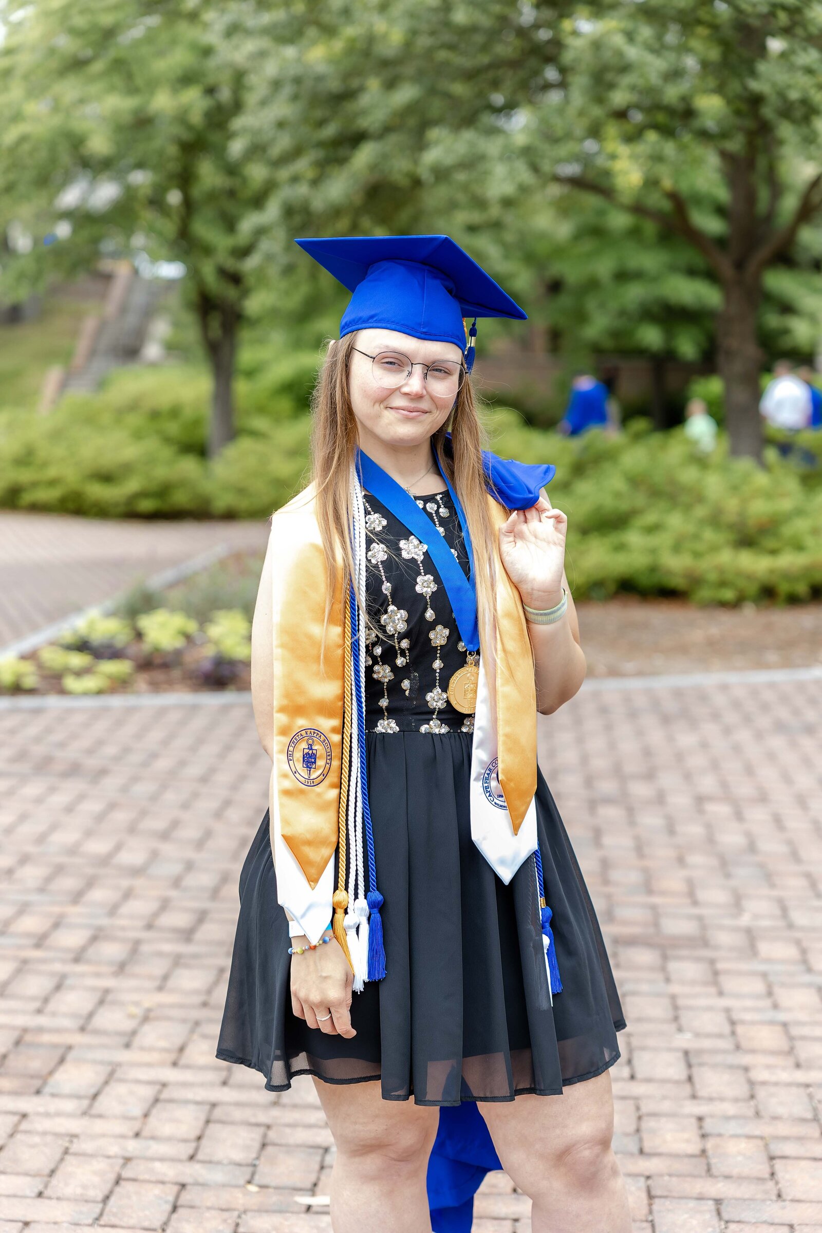 A senior proudly wearing a blue cap and gown with honor cords, standing on a paved path at Cape Fear Community College in Wilmington, North Carolina. This image highlights the achievements of graduation, perfect for students looking for artistic senior photography options.