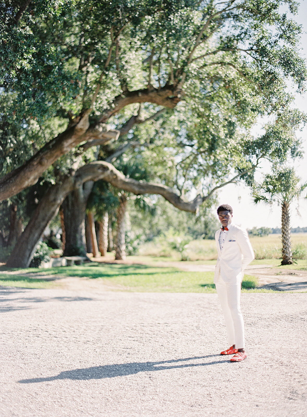 Groom in ivory and gold tuxedo with scarlet red shoes and red feather bowtie standing on the beach of Lowndes Grove. He is in the direct sunlight and his full shadow is in view, oak trees in the back of the photo. Photographed at Lowndes Grove wedding by wedding photographers in Charleston Amy Mulder Photography.