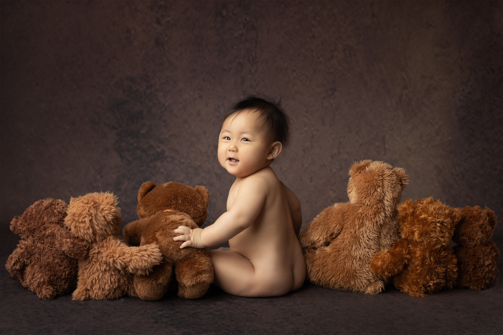 Smiling baby sits among a circle of soft brown teddy bears, looking over their shoulder with a sweet expression. A beautiful fine art milestone photo by DFW Baby Milestone Photographer Lindsay Walden.