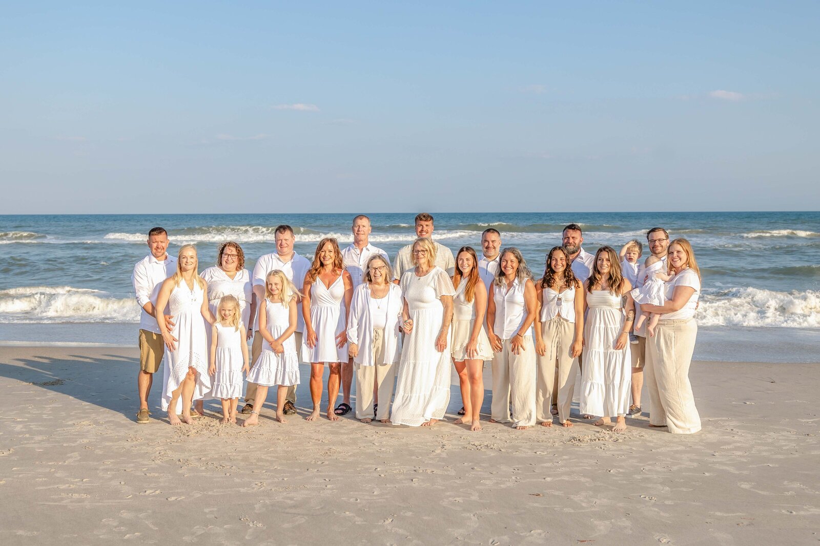A large family stands together on a sunny beach, dressed in coordinated white outfits. The bright ocean waves provide a lively backdrop, perfect for artistic family photography in Wilmington, NC, capturing vibrant and joyful family moments.
