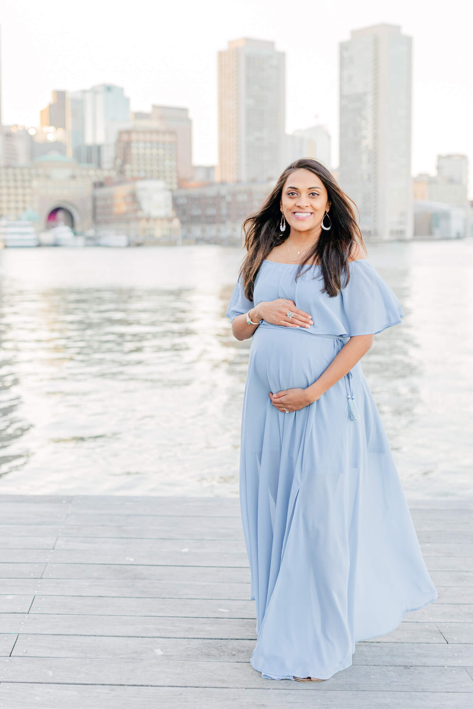 Pregnant woman holds her baby bump and smiles in front of a Boston skyline at Boston Seaport