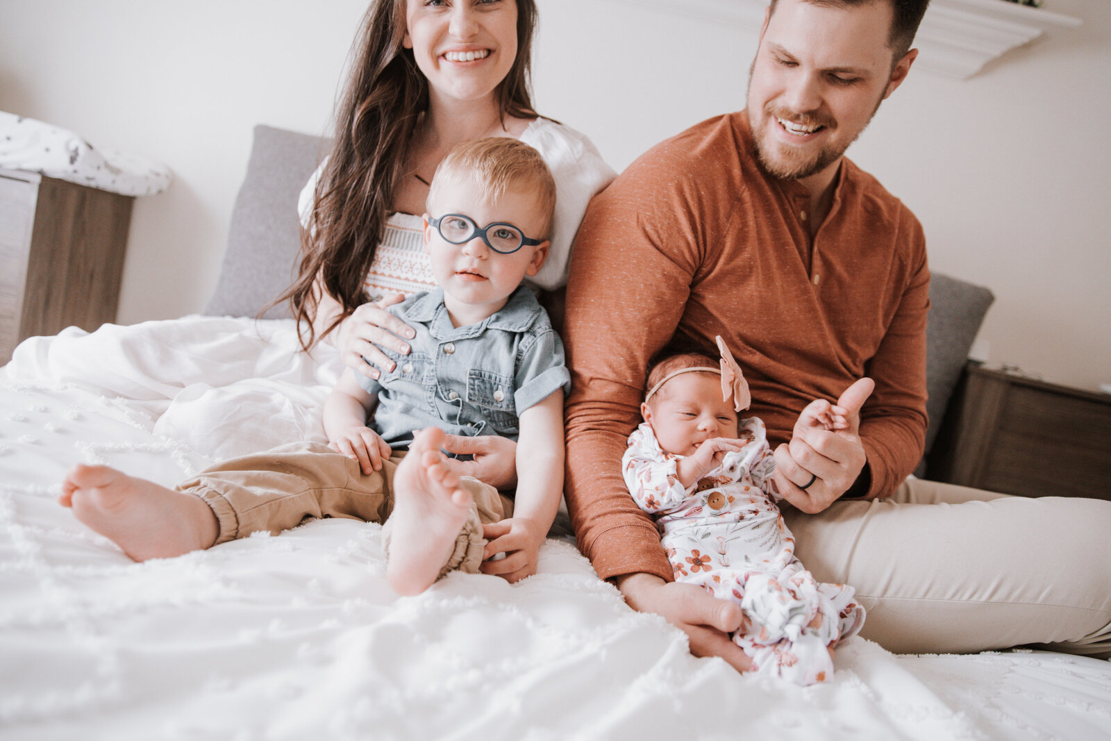 young family sitting ona bed together with mother holding toddler boy and father holding newborn girl as they all smile together for their Idaho Falls newborn photos