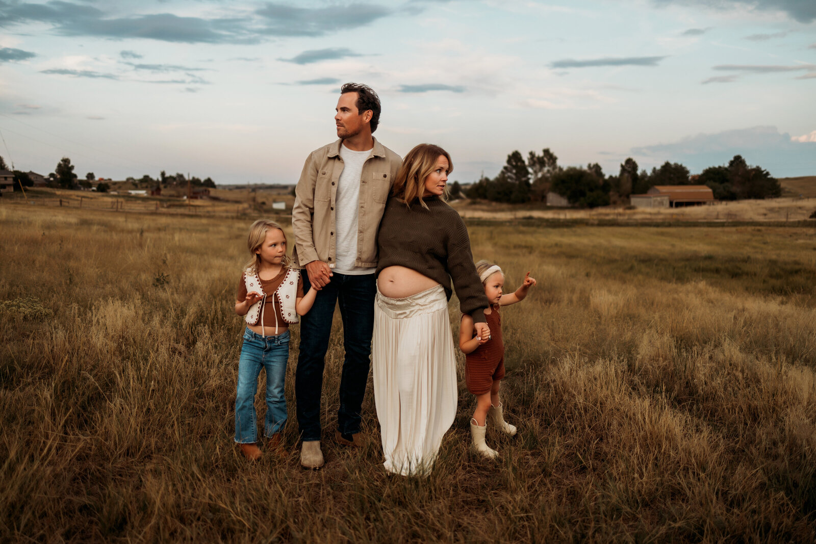 Family of four all holding hands in a field of grass while moms belly is showing off her new growing belly