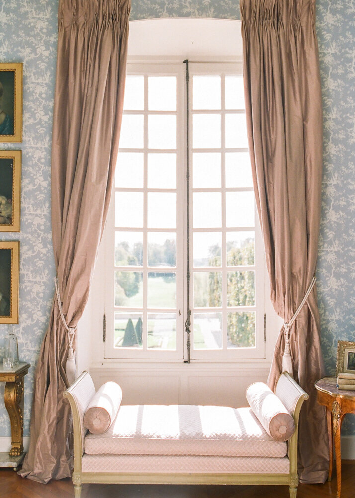 Light pouring in through large windows and a pink chaise lounge framed by beige satin curtains