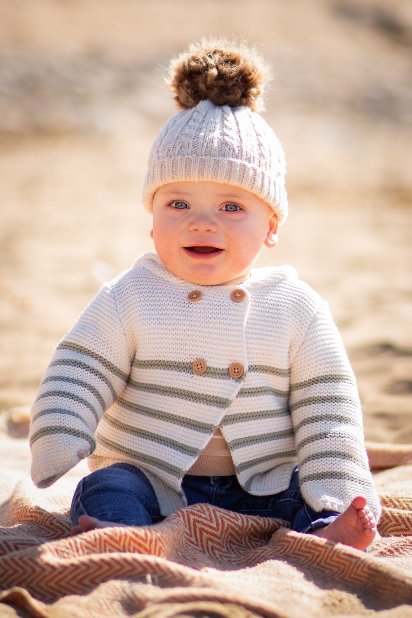 Baby sits on a blanket at the beach smiling at the camera