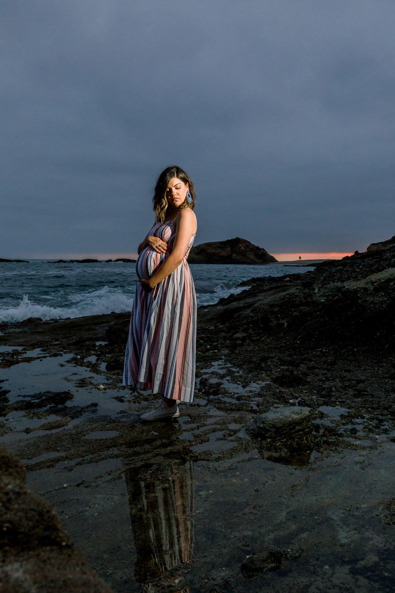 Mom to be poses on the rocks at  the  beach at night holding her belly