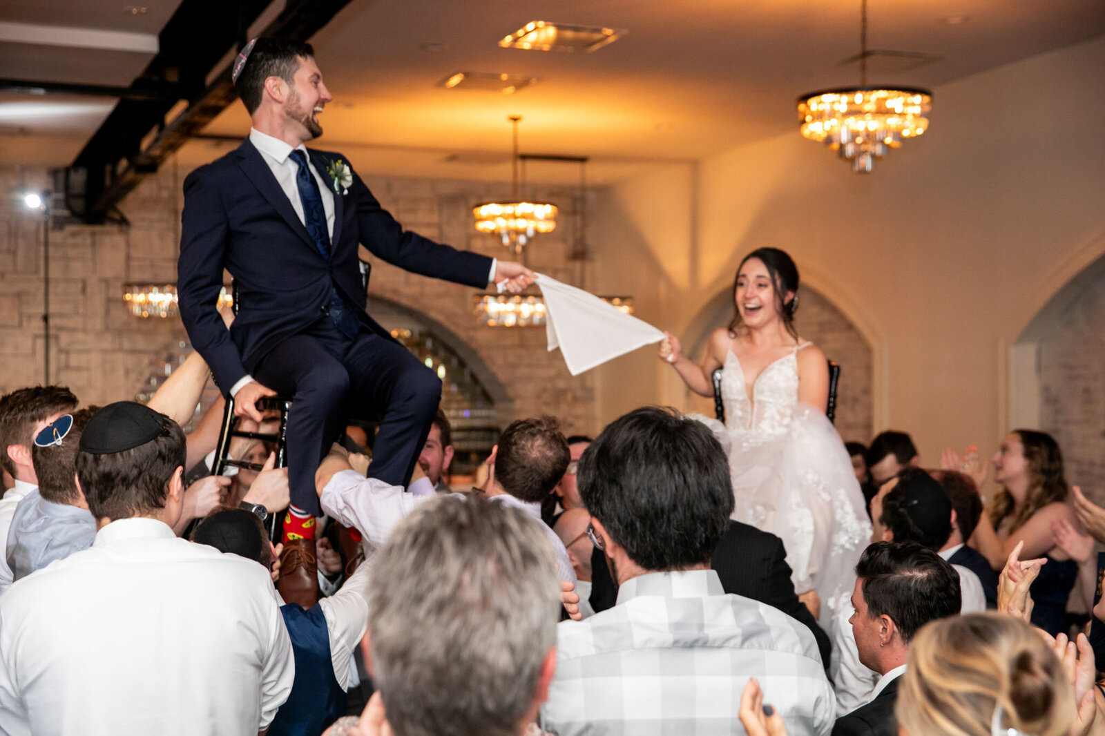 A bride and groom are lifted on chairs by guests during a lively wedding dance, holding a white cloth between them and smiling joyfully in an elegant wedding venue in st. louis