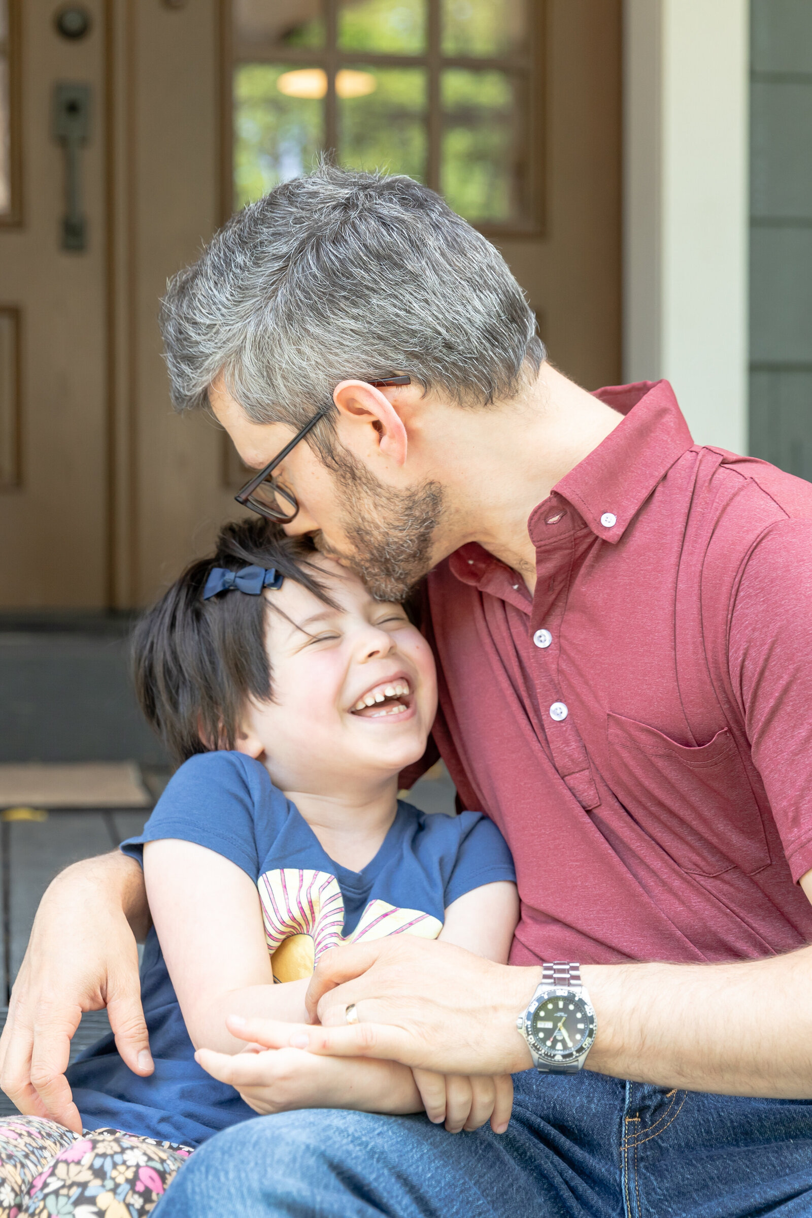 father sits with daughter and kisses her on the forehead as she laughs on porch