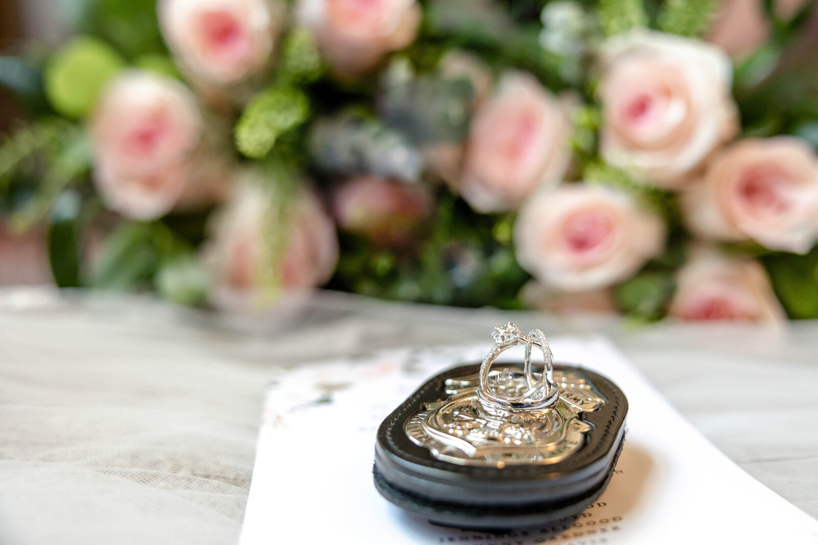 A close-up of wedding rings displayed on a black ring holder with a pink rose bouquet in the background, captured during a Wilmington, NC wedding, showcasing elegant and romantic wedding details.