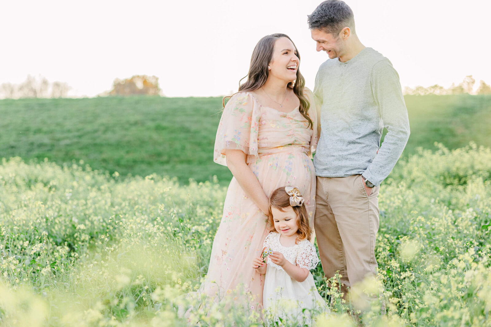 Pregnant woman, her husband, and her daughter laugh together in a field of yellow wildflowers
