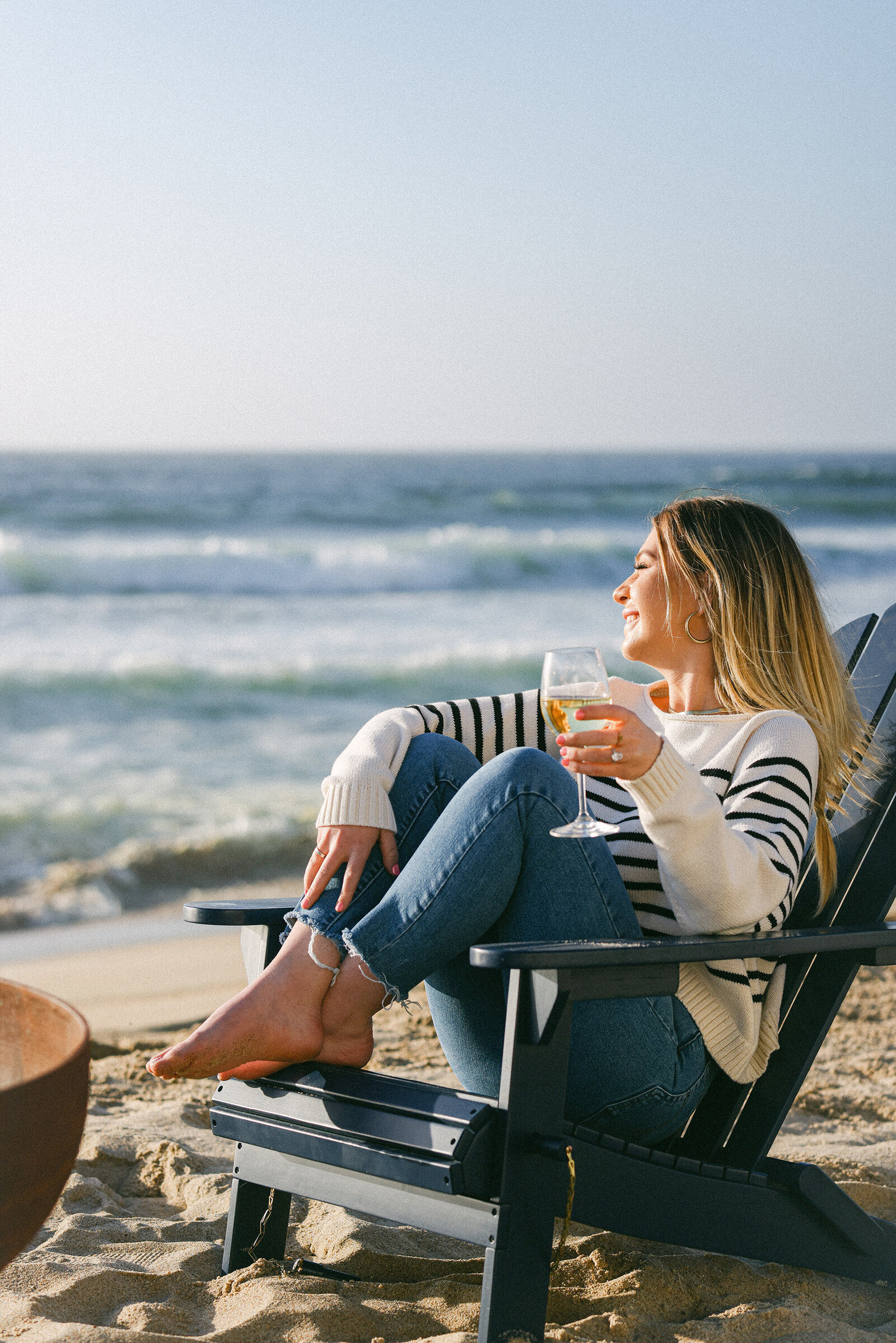 girl sitting on beach in Adirondack chair drinking wine next to bonfire in striped sweater. Luxury california monterey beach lifestyle photographer