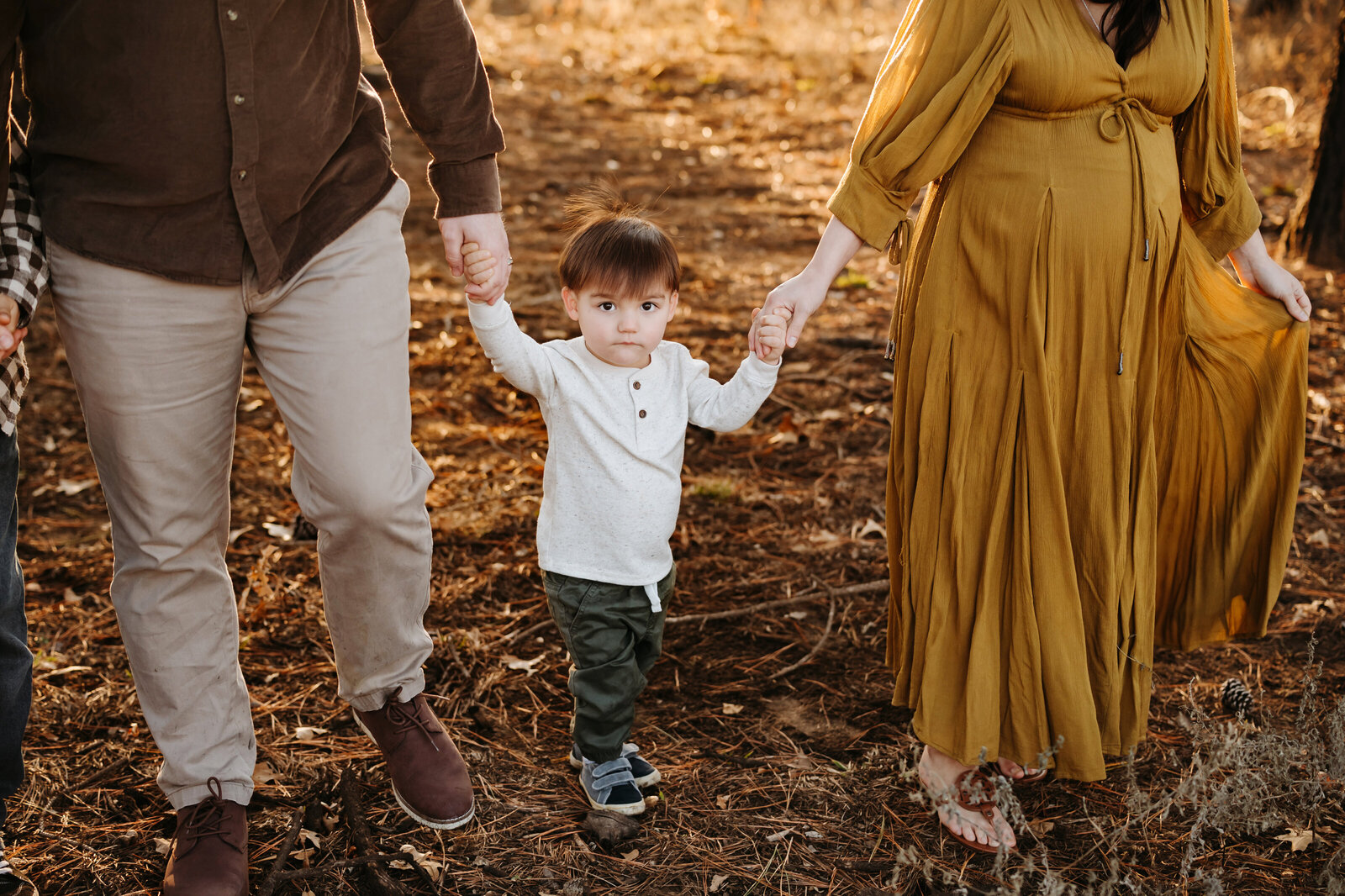 Family of three holding hands while Paula Goforth Photography captures their family session in Decatur at the Pines. Mom is wearing a long mustard color dress while Dad and son are dressed casually. 