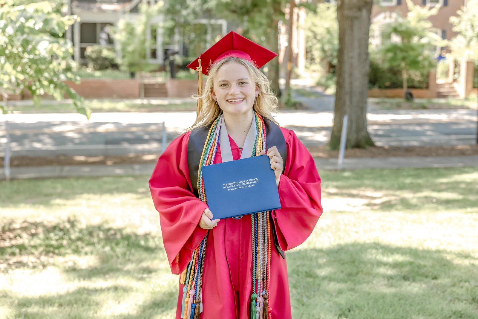 A NCSSM senior in a red graduation gown with honor cords holding a diploma in a park setting in Durham, North Carolina. This celebratory image captures the joy of academic success, perfect for meaningful senior portraits.