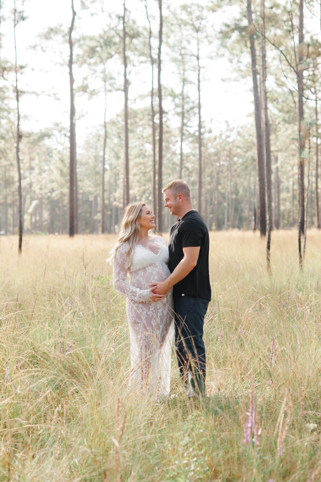 New parents stand in between tall trees and grass holding moms belly and staring deeply into each others eyes
