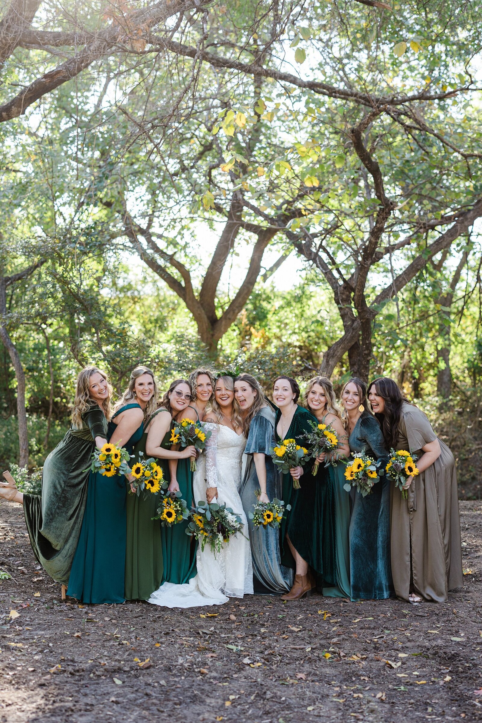 A bride posing very closely with all of her bridesmaids in a wooded setting where her ceremony took place. The bride is wearing a long white dress and a colorful flower crown while holding a bouquet. The bridesmaids are all wearing dresses of varying shades of blue and green while holding bouquets of sunflowers.