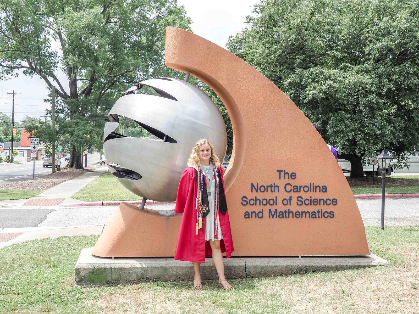 A graduating senior in a red graduation gown standing by the North Carolina School of Science and Mathematics sign. This image celebrates academic excellence, perfect for capturing meaningful and professional senior portraits.