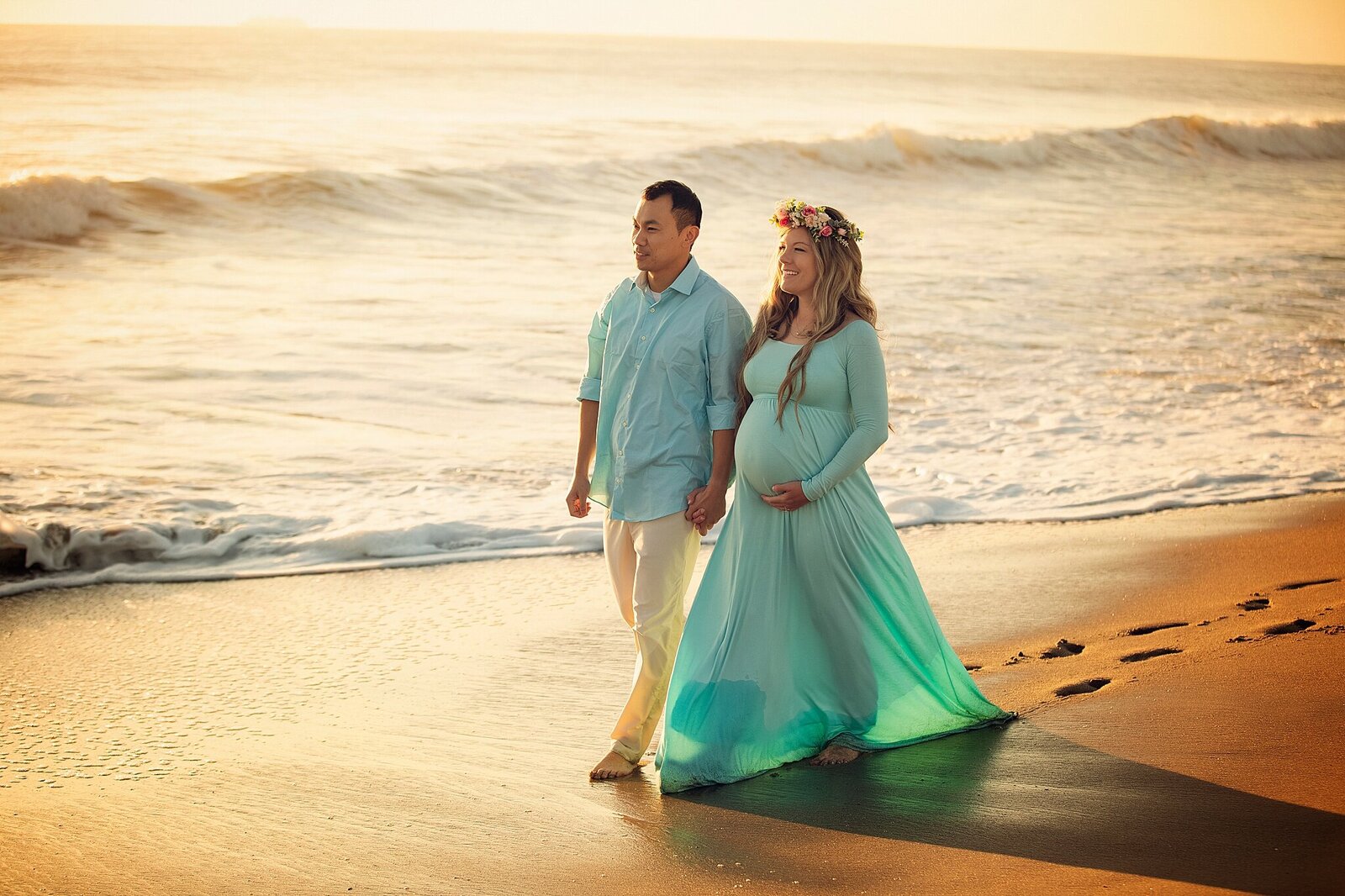 A pregnant couple strolls hand-in-hand along the beach during a maternity photography session by Bia Schaefer.