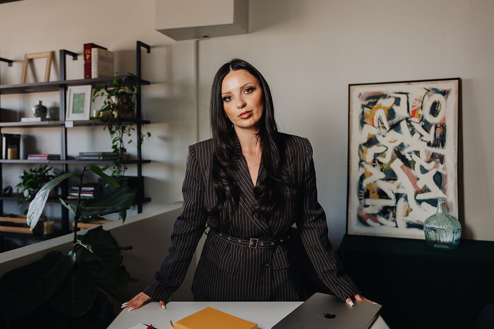 Woman in pinstripe suit standing at desk with laptop in styled office, abstract art behind.