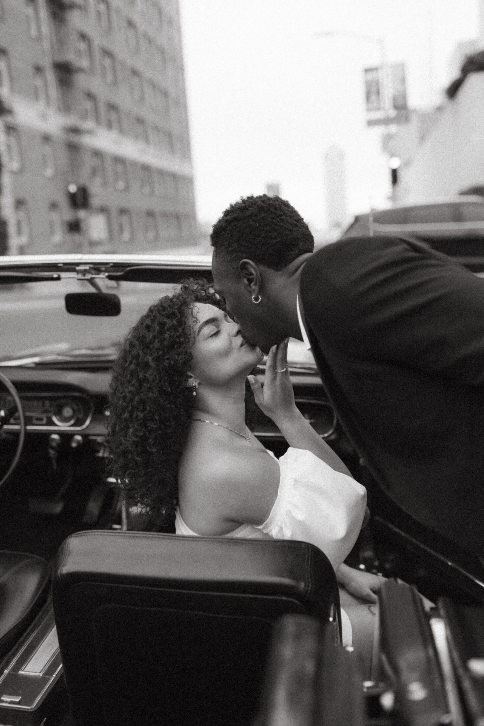 A monochrome photo of a man kissing a woman as they sit in a vintage car in San Francisco, conveying a tender moment between the couple.