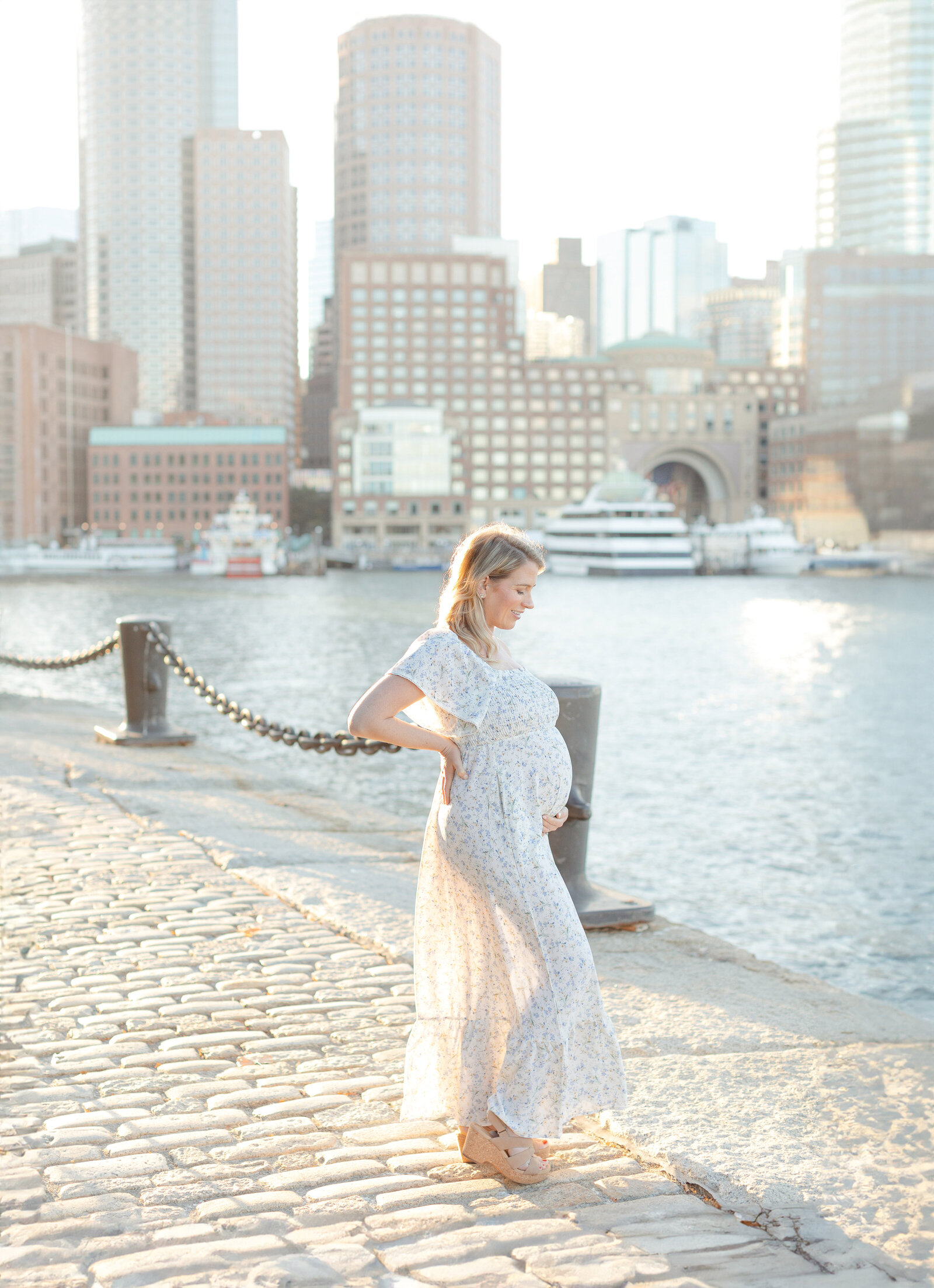 woman wearing flowy, floral dress holds baby bump during sunset boston seaport photo session