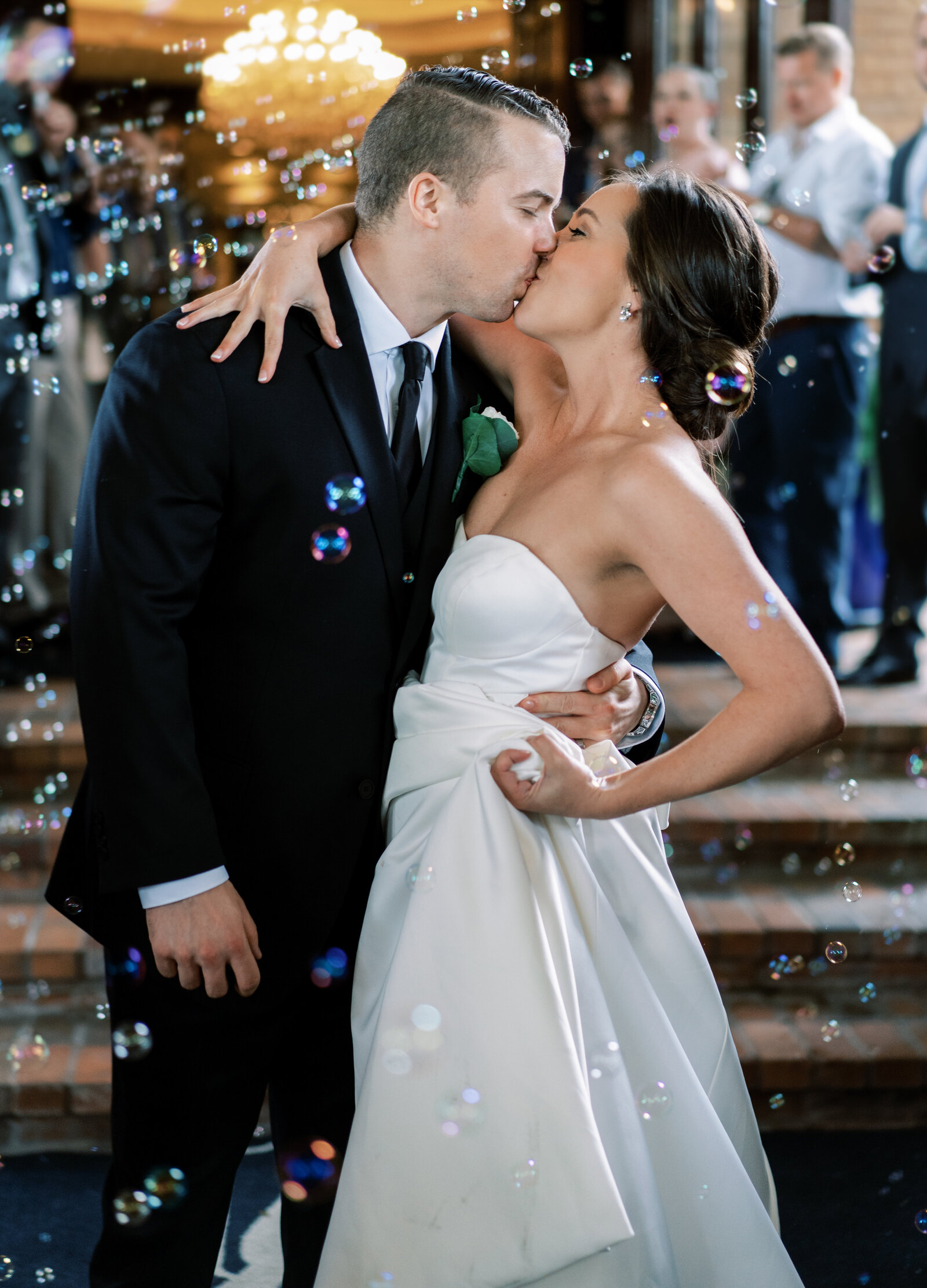 Portrait of a bride and groom kissing while exiting a building with bubbles and a crowd in the back.