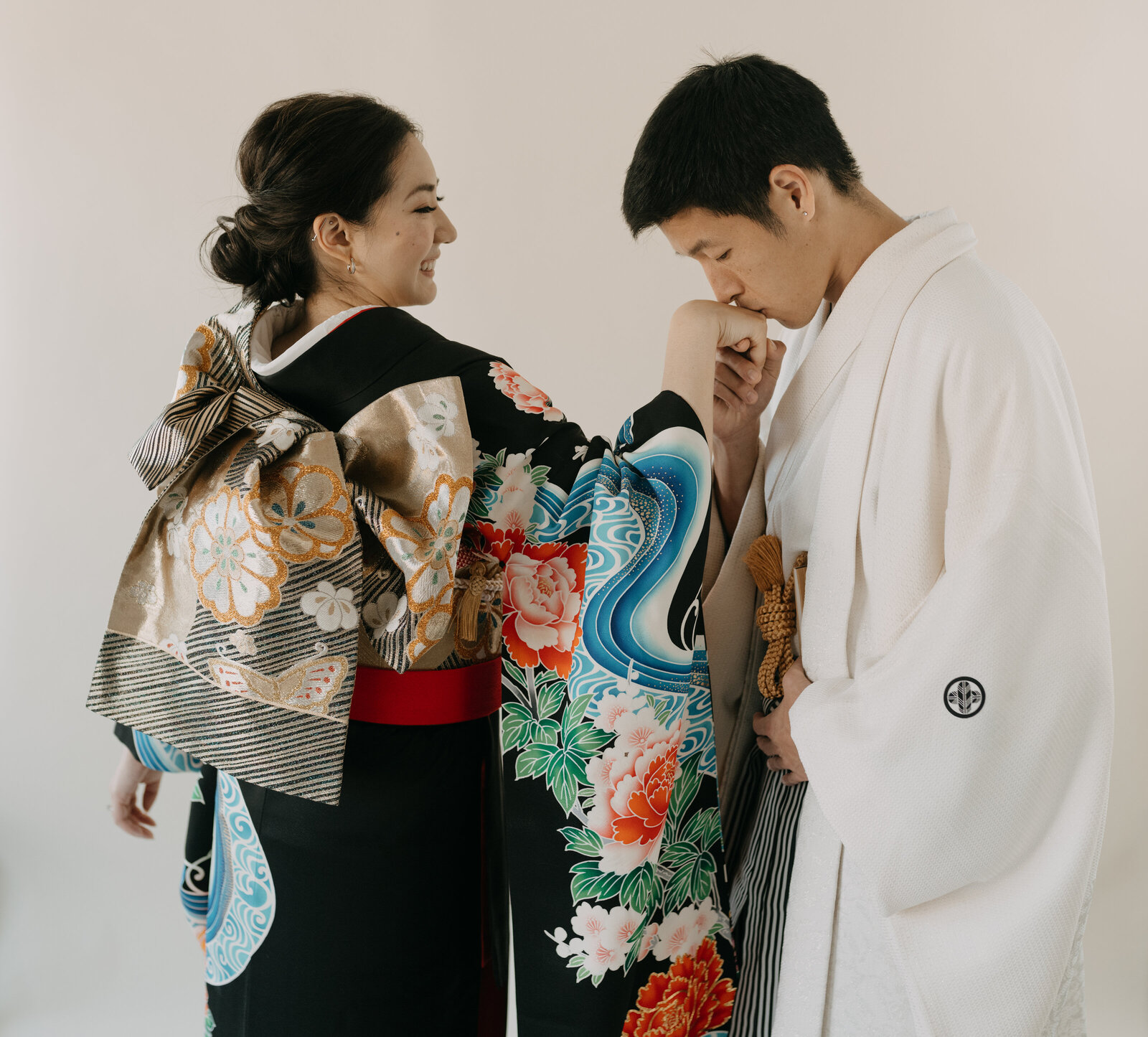 Bride in elegant black wedding kimono rental with vivid peony design, groom in white montsuki gently kissing her hand, Los Angeles California studio session | 黒引き振袖レンタル 豪華牡丹柄 新郎が花嫁の手にキスするスタジオフォト