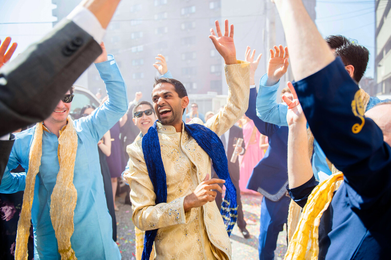 A joyful man in a traditional gold and blue outfit dances amidst a lively group of people. The scene is outdoors during the day, filled with vibrant energy and happiness as everyone celebrates, some in colorful attire and sunglasses.